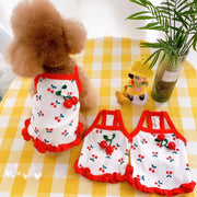 Dog wearing a Cherry Dress for Dogs on a yellow checkered tablecloth with two smaller dresses and a toy.
