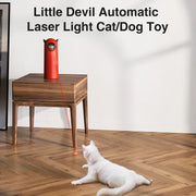 Red Little Devil Automatic Infrared LED Laser Cat Toy cat/dog toy on a wooden table with a white cat playing with it on a wooden floor.