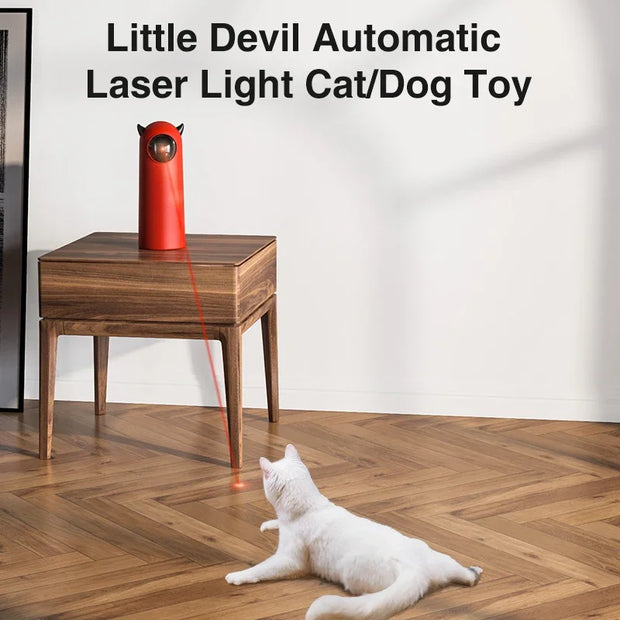 Red Little Devil Automatic Infrared LED Laser Cat Toy cat/dog toy on a wooden table with a white cat playing with it on a wooden floor.