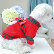 Small dog wearing a red Christmas Dog Sweater with green stripes and a bow, sitting on a white surface with flowers in the background.
