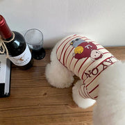 Small dog wrapped in a Striped Bear T-Shirt with a bear design on a wooden surface next to a bottle of wine and a glass.