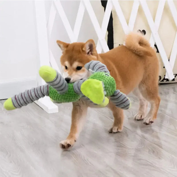 Dog playing with a green and gray Plush Animal Dog Toys on a light wooden floor.