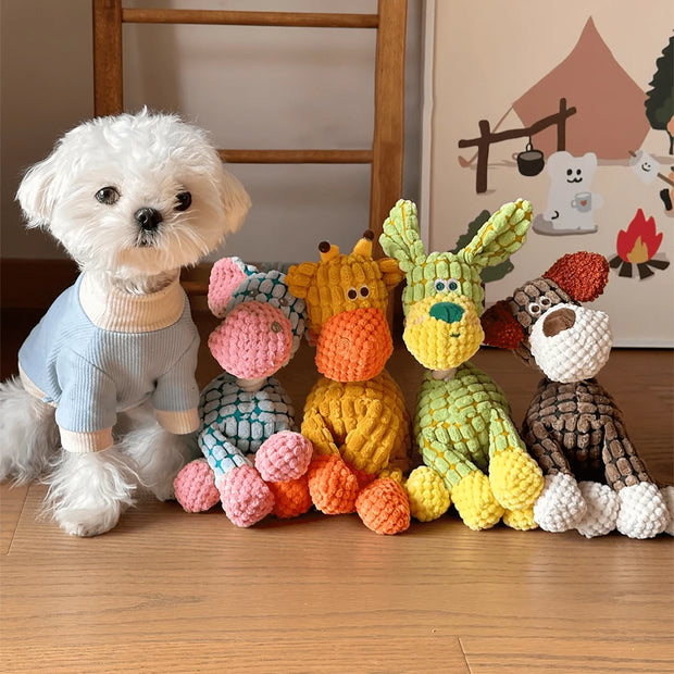 White dog wearing a blue sweater standing next to colorful knitted Plush Animal Rope Dog Toys on a wooden floor.