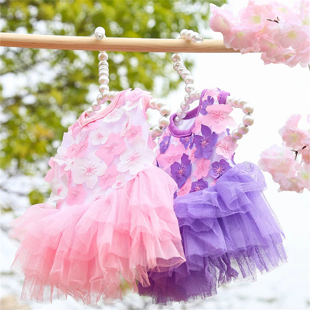 Two Adorable Floral Dog Dress, one pink and one purple, hanging on a rack with cherry blossoms in the background.